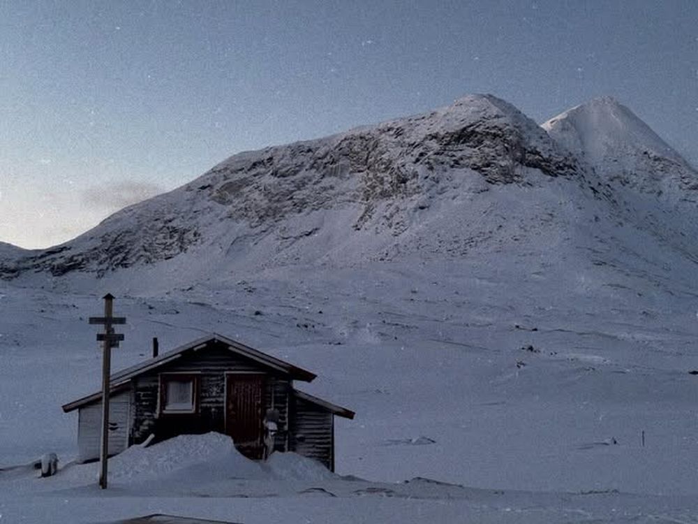 Liten fjellstue foran et snødekt fjell under stjernehimmel.